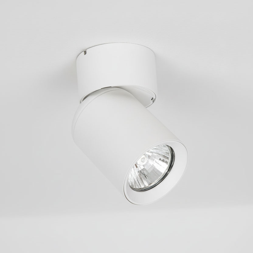 Biała lampa sufitowa typu spot, montowana na suficie, w kształcie cylindrycznym. Kąt ujęcia z boku, ukazujący detal reflektora. Prosta konstrukcja, idealna do nowoczesnych wnętrz.