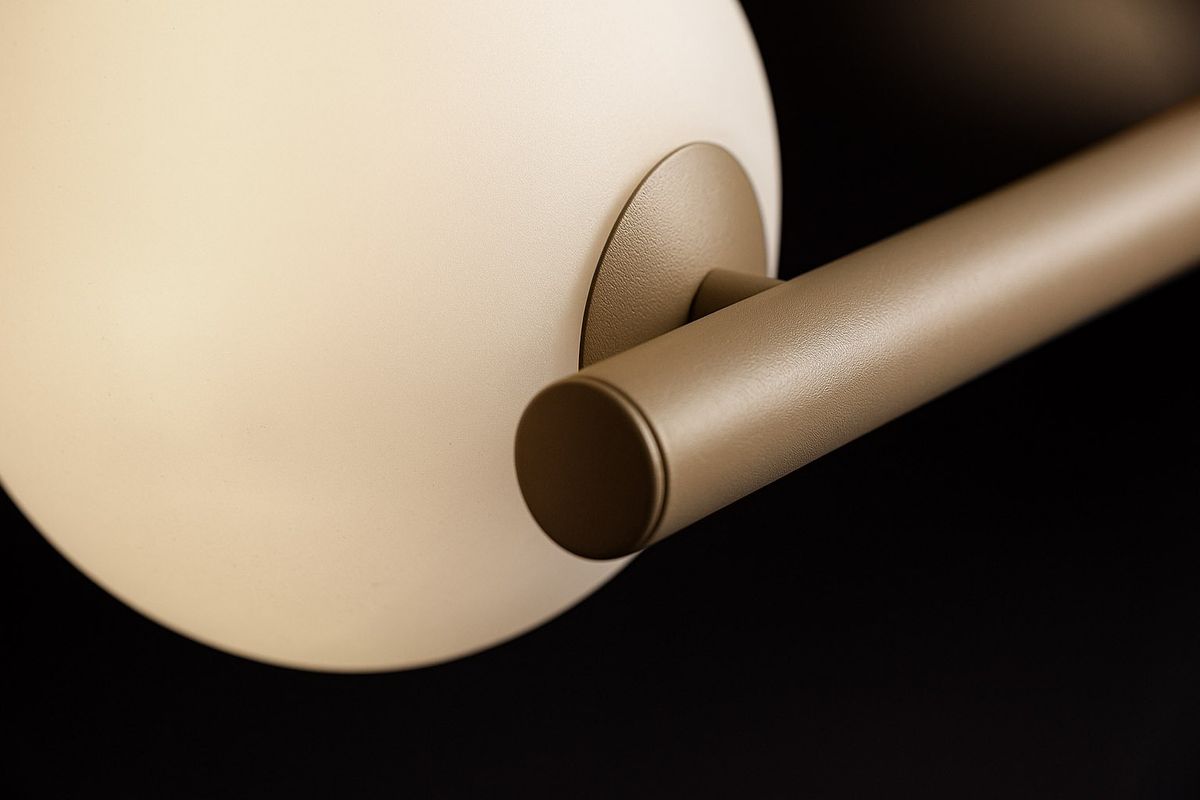 Beżowa lampa wisząca Ardi K-5842 z siedmioma gniazdami G9. Zbliżenie na metalowy uchwyt oraz okrągły klosz, wykonany z matowego materiału. Prosta, elegancka konstrukcja, montowana na suficie. Tło ciemne, co podkreśla kolor lampy.