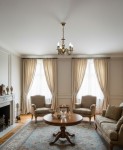 Żyrandol świecznikowy Antares w odcieniu patyny, z czterema miejscami na żarówki E14, zawieszony na delikatnym łańcuchu. Lampa umieszczona w eleganckim wnętrzu z sofą, fotelami i oknami z zasłonami.