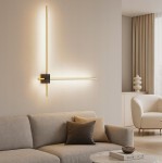 Kinkiet LED 12W 4000K w kolorze złotym i czarnym, zamontowany na ścianie. Składa się z dwóch prostokątnych elementów świetlnych w formie linii. Otoczenie to nowoczesny salon z sofą i stolikiem. Tło w jasnym kolorze.