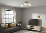 Lampa sufitowa Bali II z trzema oprawami E27. Złota metalowa konstrukcja z okrągłymi, białymi kloszami. Ujęcie z boku w nowoczesnym wnętrzu z szarymi ścianami i sofą, na której leżą poduszki w odcieniach złota.