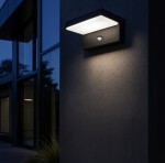 Hestio LED kinkiet elewacyjny w kolorze czarnym, zamontowany na ścianie, z prostokątną, białą osłoną lampy i wbudowanym czujnikiem ruchu. Oświetlenie emituje delikatne światło, w tle widoczna elewacja budynku w ciemnym otoczeniu.
