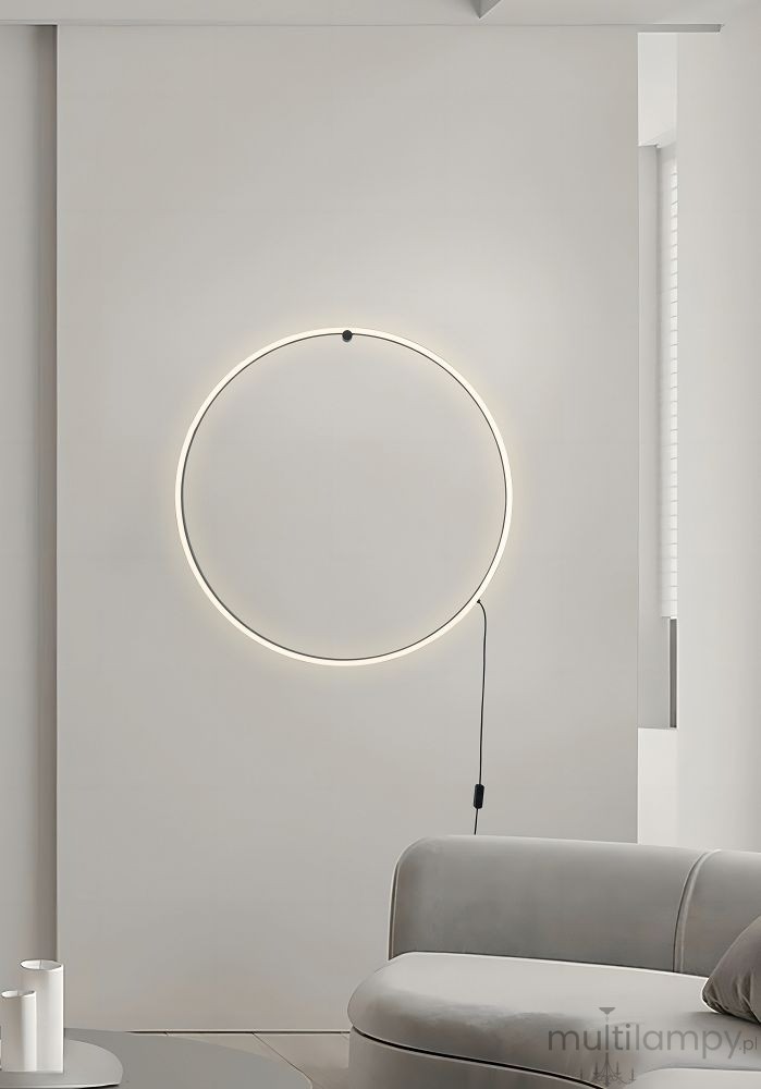 Kinkiet Scopello 1xLED/20W CCT w kolorze czarnym, w kształcie okręgu, zamontowany na jasnej ścianie. Widziany z przodu, z kablem oprawy widocznym po prawej stronie. Obok widoczna szara sofa i okrągły stół z białymi świecznikami.