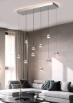 Lampa wisząca Quiberon z 10 szklanymi kloszami LED, zawieszona na cienkich stalowych linkach, w wykończeniu niklowym. Umieszczona na prostokątnej podstawie, w nowoczesnym wnętrzu z szarymi ścianami i sofą.