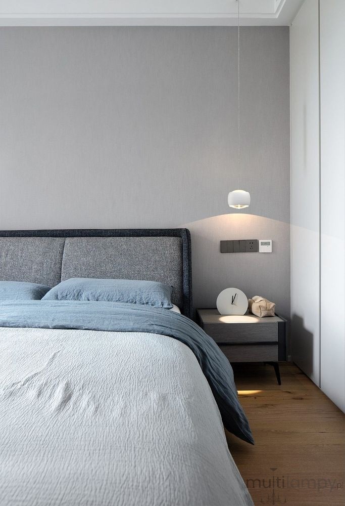 Lampa wisząca Quiberon 1xLED 8W w kolorze białym, zawieszona nad łóżkiem, z okrągłym kloszem. W tle widoczna szara ściana oraz nowoczesne meble. Oświetlenie delikatnie podkreśla aranżację sypialni.