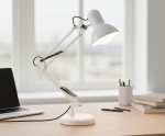 Biała lampka biurkowa Lena z podstawą i uchwytem do stołu, z regulowanym ramieniem. Ujęcie z boku, na drewnianym biurku obok laptopa i przyborów biurowych. Widoczne sprężyny na ramieniu oraz okrągła podstawa.
