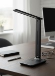 Czarna lampka biurkowa Kivo LED o mocy 5W, z regulowanym ramieniem i prostokątną podstawą. W tle widoczne akcesoria biurowe, takie jak notes, długopis i okulary. Produkt ustawiony na drewnianym biurku, w jasnym wnętrzu.