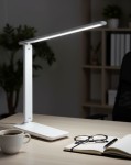 Kivo LED lampka biurkowa 5W/CCT w kolorze białym, z regulowanym ramieniem, ustawiona na biurku. W tle widać rośliny i półki. Obok lampki filiżanka kawy, okulary oraz notes z długopisem.