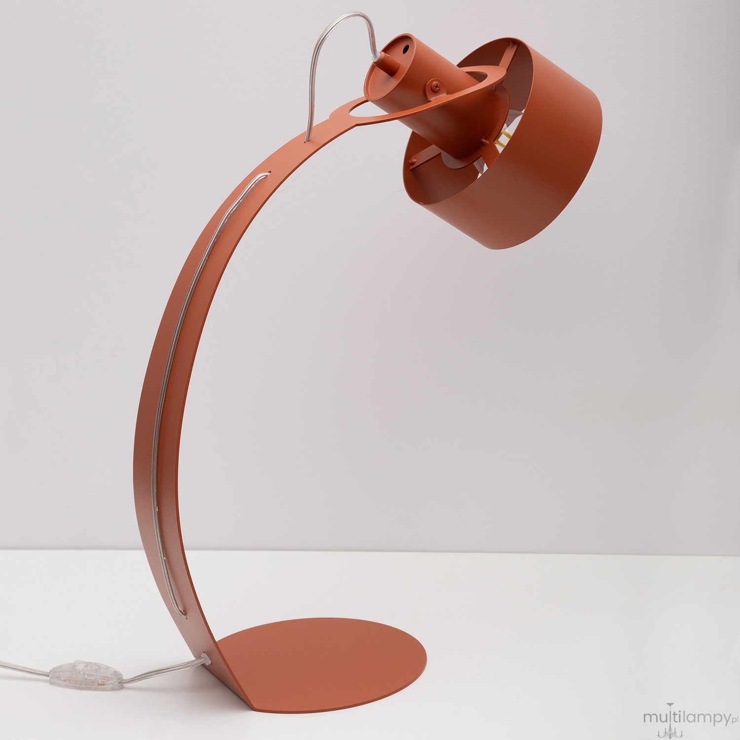 Lampa stołowa Rif w kolorze ceglastym z metalowym kloszem na żarówkę E27. Posiada smukłą, zakrzywioną podstawę oraz przewód z włącznikiem, ustawiona na białym tle, w pozycji stojącej.