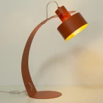 Lampa stołowa Rif w kolorze ceglastym z metalowym, okrągłym kloszem i giętką podstawą. Ujęcie z boku, lampka skierowana w dół. Widoczny przewód z przełącznikiem. Wysoka, nowoczesna forma, idealna do biura lub na stół.