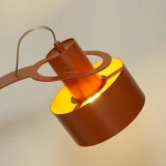 Lampa stołowa Rif w ceglanym kolorze z oprawą na żarówkę E27. Ujęcie z góry, ukazujące okrągły klosz i metalowy element mocujący. Wnętrze klosza w kolorze żółtym, co podkreśla ciepłe światło. Kabel zasilający widoczny z tyłu lampy.