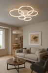 Lampa sufitowa Ravello z pięcioma okrągłymi pierścieniami LED w kolorze białym, zamontowana na suficie. Okręgi emitują ciepłe światło, tworząc nowoczesny akcent w aranżacji wnętrza. Tło to stylowy salon z sofą i stołem kawowym.