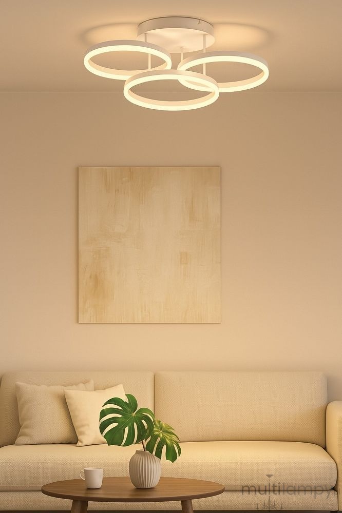 Lampa sufitowa Ravello 3xLED 48W w kolorze piaskowym z trzema okrągłymi pierścieniami emitującymi ciepłe światło. Zawieszona na płaskim, okrągłym stelażu. Widoczne tło w neutralnych barwach, z kanapą i rośliną w donicy.