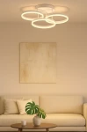 Lampa sufitowa Ravello 3xLED 48W w kolorze piaskowym z trzema okrągłymi pierścieniami emitującymi ciepłe światło. Zawieszona na płaskim, okrągłym stelażu. Widoczne tło w neutralnych barwach, z kanapą i rośliną w donicy.