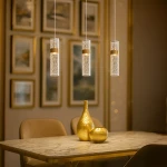 Lagos lampa wisząca LED 8W 4000K w złotej oprawie, o cylindrycznym kształcie z delikatnym wzorem, zawieszona na cienkich linkach. W tle widoczna elegancka aranżacja z obrazami oraz złotymi dekoracjami na stole.