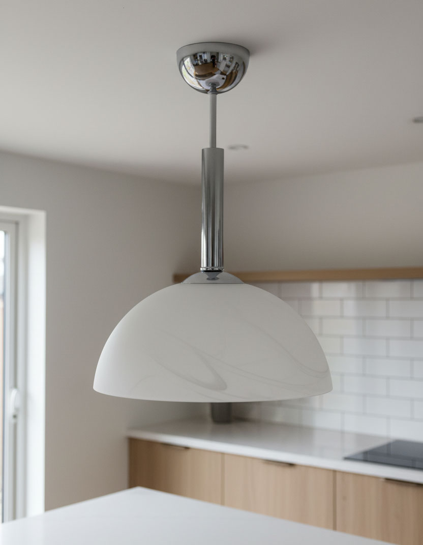 Lampa wisząca Klara z chromowanym wykończeniem i białym kloszem w kształcie półkuli, zawieszona na cienkiej metalowej rurze. Widoczna w nowoczesnej kuchni, z jasnym tłem i drewnianymi elementami mebli.