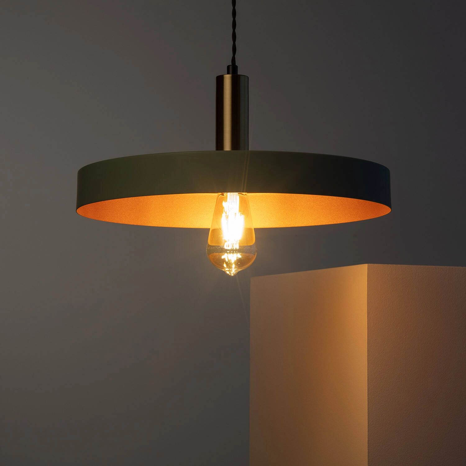 Lampa wisząca Dario w kolorze szałwiowym z złotym wnętrzem, z oprawą na żarówkę E27. Ujęcie z boku, zwisająca z sufitu, z widoczną żarówką w szklanej obudowie, emitującą ciepłe światło na sąsiednią ścianę.