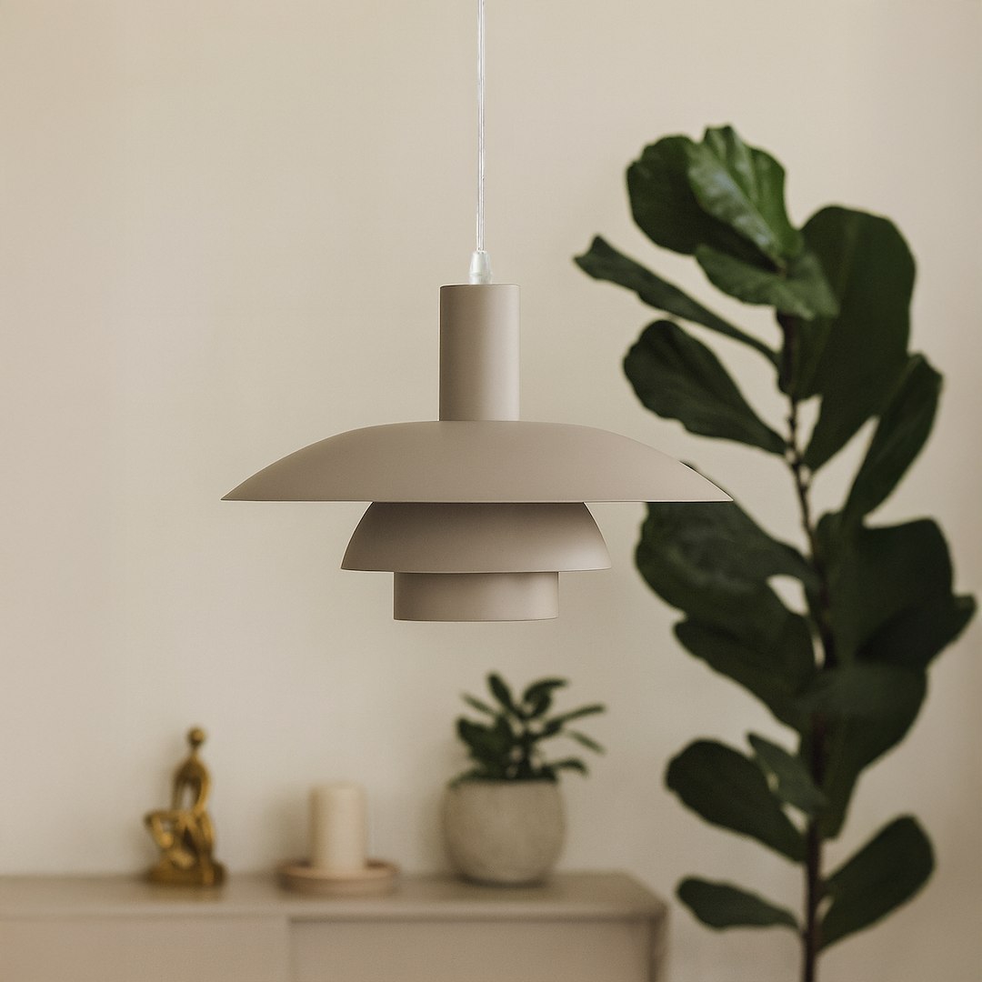 Lampa wisząca Ferro w kolorze kaszmirowym, z metalowym kloszem składającym się z trzech poziomych elementów, zawieszona na przezroczystym przewodzie. W tle roślina doniczkowa i minimalistyczne dekoracje.