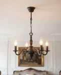 Żyrandol świecznikowy Cereus w kolorze czarnym, z czterema oprawami E14. Ma klasyczny kształt z ozdobnymi ramionami i eleganckimi żarówkami w kształcie łzy. Zawieszony na łańcuchu, w aranżacji z jasnym tłem i eleganckim meblem.