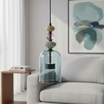 Lampa wisząca Arte z kloszem w kształcie dzwonu, wykonana z przezroczystego, niebieskiego szkła. U góry ozdobne elementy w kolorze czarnym, beżowym i różowym. Zawieszona na czarnym przewodzie, w aranżacji z szarym sofą i abstrakcyjnym obrazem w tle.