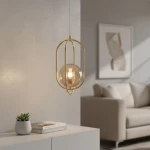 Lampa wisząca Binar 1-punktowa w kolorze złotym, z przezroczystą kulistą żarówką. Ujęcie z boku, zawieszona na białym przewodzie, w nowoczesnym wnętrzu z jasnym tłem i kanapą. Widoczna roślinka w doniczce oraz stos książek na białym meblu.