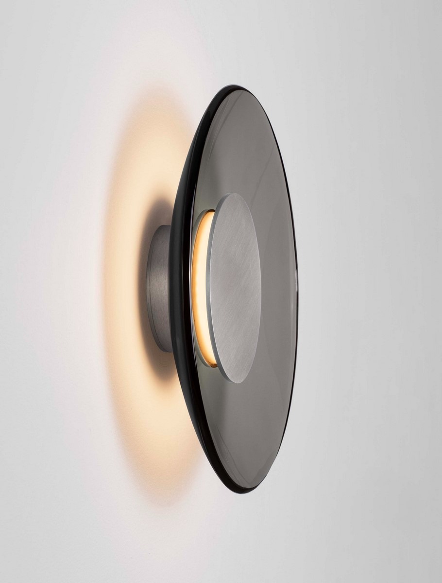 Kinkiet Huanuco 1xLED 17W w kolorze niklowym z aluminium. Okrągła lampa z przezroczystym kloszem, emitująca ciepłe światło, zamontowana na ścianie. Widoczny centralny element w kolorze matowego niklu. Tło jasne.