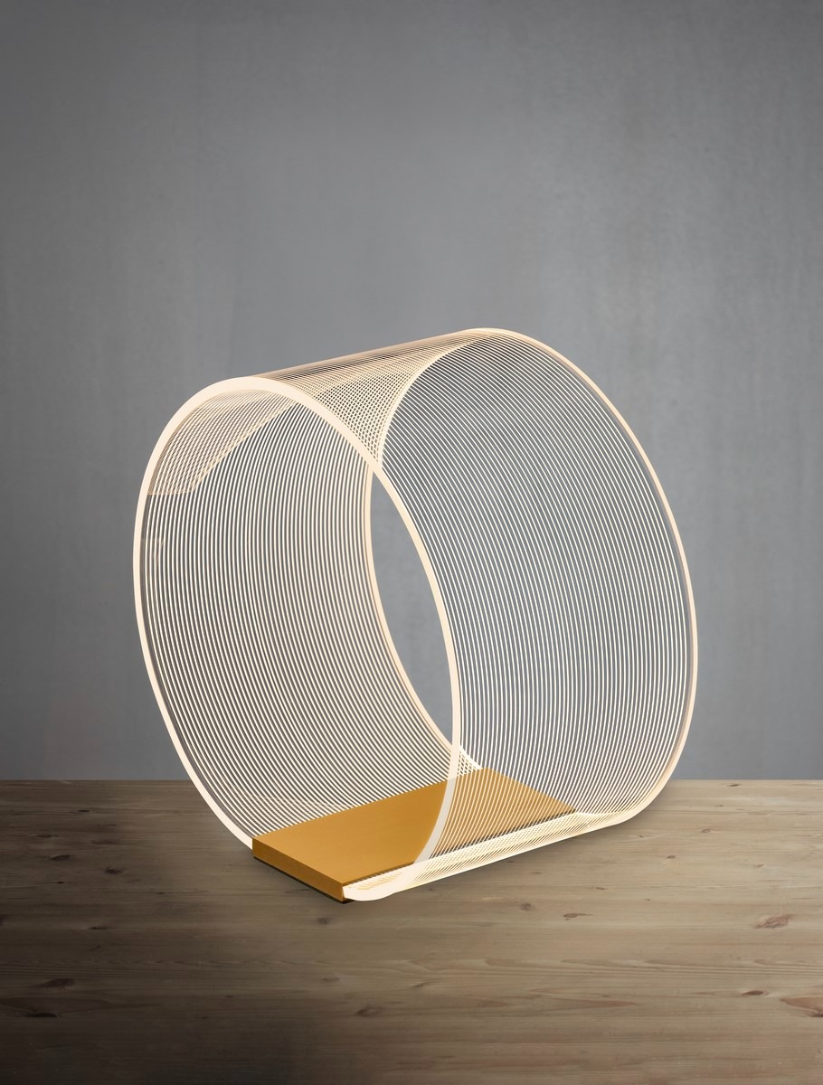 Lampa stołowa Huaral z jedną diodą LED 14W, o kształcie okręgu. Złota podstawa z gładkiego metalu, a klosz wykonany z cienkich, spiralnych linii, emitujących miękkie światło. Produkt ustawiony na drewnianym blacie, z szarym tłem.