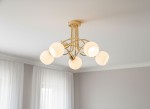 Lampa wisząca Eryka z pięcioma źródłami światła E14, złota konstrukcja z metalowymi ramionami. Okrągłe, białe klosze z matowego szkła. Ujęcie z dołu, lampa zamontowana na suficie, w tle jasne zasłony.