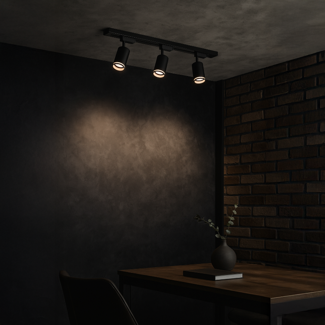 Zestaw szynowy 1m Pipe Ring Track z trzema reflektorami GU10 w kolorze czarnym, zamontowany na ścianie. Reflektory skierowane w dół, na tle ciemnej ściany z teksturowanym wykończeniem, obok drewnianego stołu z dekoracjami.