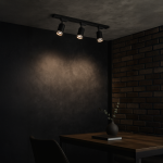 Zestaw szynowy 1m Pipe Ring Track z trzema reflektorami GU10 w kolorze czarnym, zamontowany na ścianie. Reflektory skierowane w dół, na tle ciemnej ściany z teksturowanym wykończeniem, obok drewnianego stołu z dekoracjami.