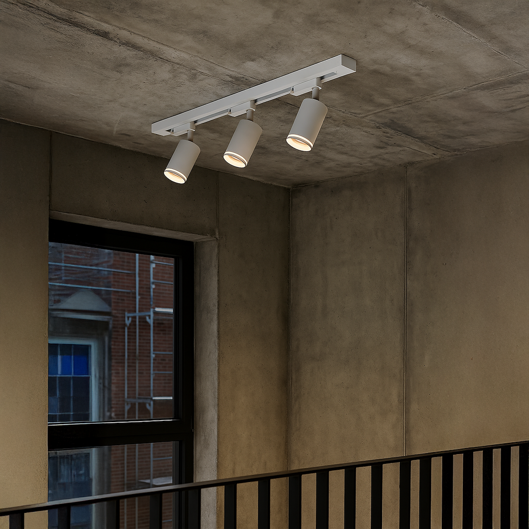 Zestaw szynowy 1m Pipe Ring Track 3xGU10 w kolorze białym. Trzy okrągłe reflektory zamontowane na prostokątnej szynie, zawieszone pod sufitem w minimalistycznym wnętrzu z betonowymi ścianami i dużym oknem.