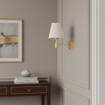 Kinkiet Astrit 1xE14 w kolorze złotym, z abażurem w odcieniu jasnego beżu. Montowany na ścianie, z widoczną podstawą okrągłą i prostym ramieniem. W tle drewniana komoda z dekoracjami.