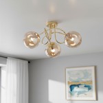 Lampa sufitowa Banko z trzema źródłami światła E14, w złotym kolorze. Kształt z delikatnymi, zaokrąglonymi kloszami w odcieniu przezroczystym. Montowana na suficie, z dekoracyjnymi, falistymi ramionami. Tło w jasnych tonacjach z obrazem na ścianie.