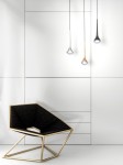 Lampa wisząca Crima Gold LED w kształcie kropli, w kolorze złotym, zawieszona na cienkim przewodzie. Oświetlenie znajduje się w minimalistycznym wnętrzu, obok nowoczesnego fotela o geometrycznej formie i czarnym obiciu. Tło białe i gładkie.