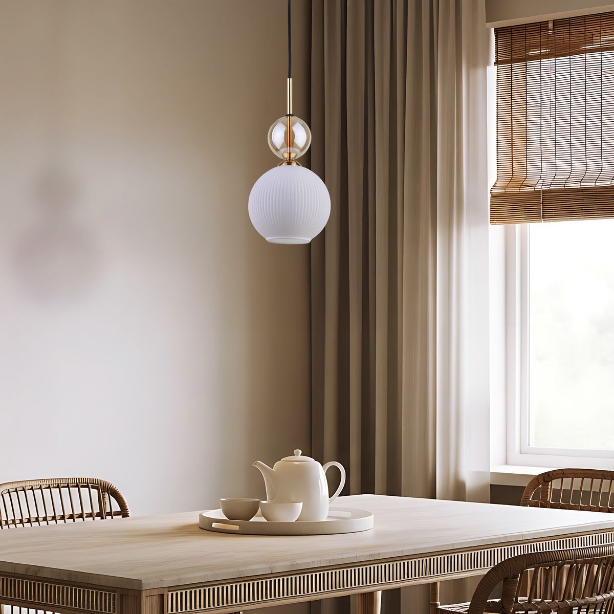 Lampa wisząca Sophia 1xE14 w kolorze białym z teksturowanym kloszem, zawieszona na cienkim czarnym przewodzie. Część metalowa w złotym kolorze, umieszczona nad okrągłym kloszem. Tło neutralne, z drewnianym stołem i filiżankami w jasnych odcieniach.