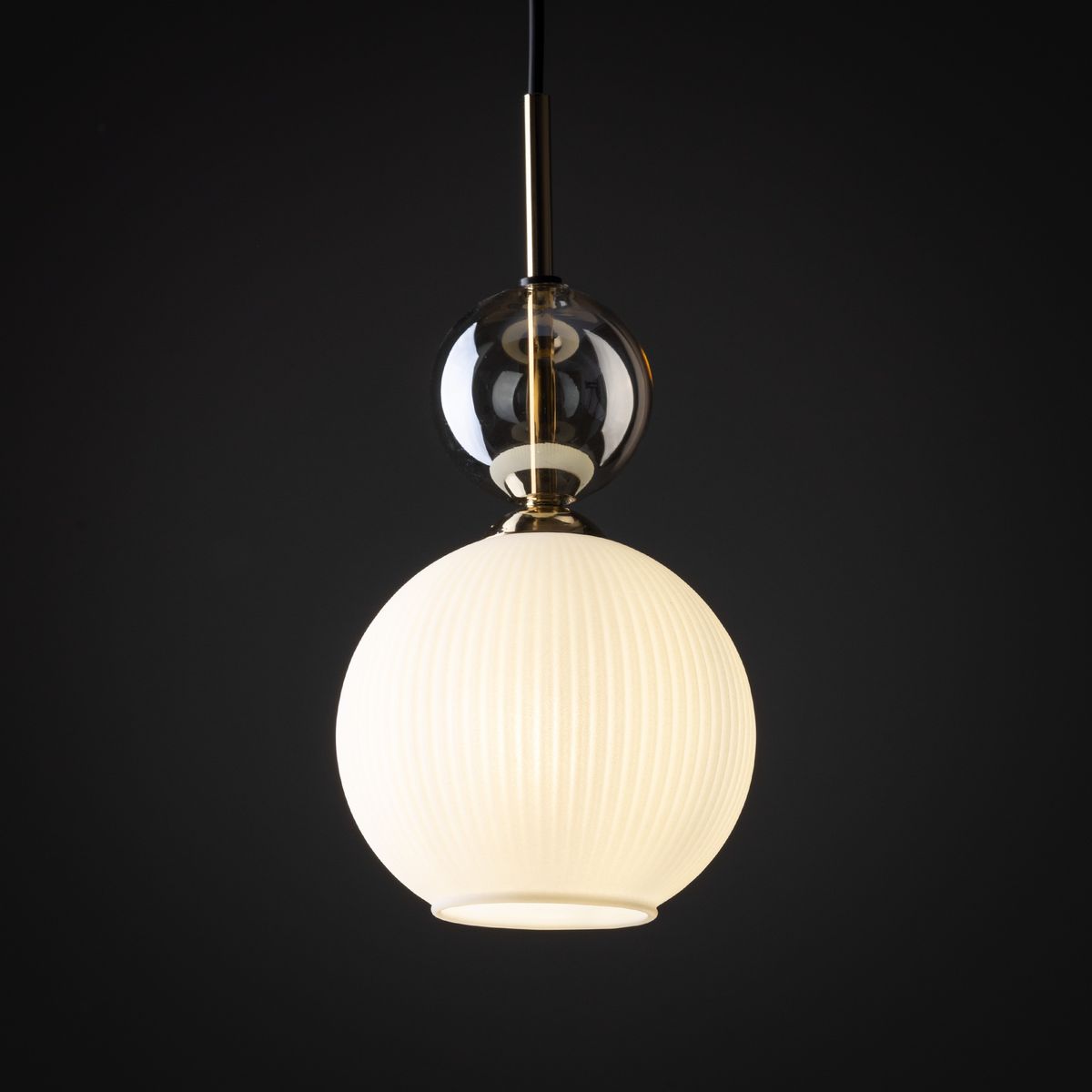 Lampa wisząca Sophia z kloszem w kształcie białej, matowej kuli z wypukłymi żłobieniami, umieszczona na złotej oprawie. Górna część z przezroczystego szkła. Ujęcie frontalne na ciemnym tle. Działa z żarówką E14.