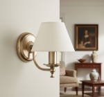 Kinkiet Sofia 1-punktowy 18340 w odcieniu złotym z białym abażurem, zamontowany na ścianie. Posiada dekoracyjną podstawę z wykończeniem w stylu klasycznym oraz uchwyt w kształcie łuku. W tle widoczny fragment eleganckiego wnętrza.