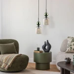 Lampa wisząca Sophia 1xE14, z bursztynowym kloszem i zieloną kulą, zawieszona na czarnym przewodzie. W tle widoczny nowoczesny salon z zielonym fotelem, okrągłym stołem i dekoracjami w neutralnych kolorach.