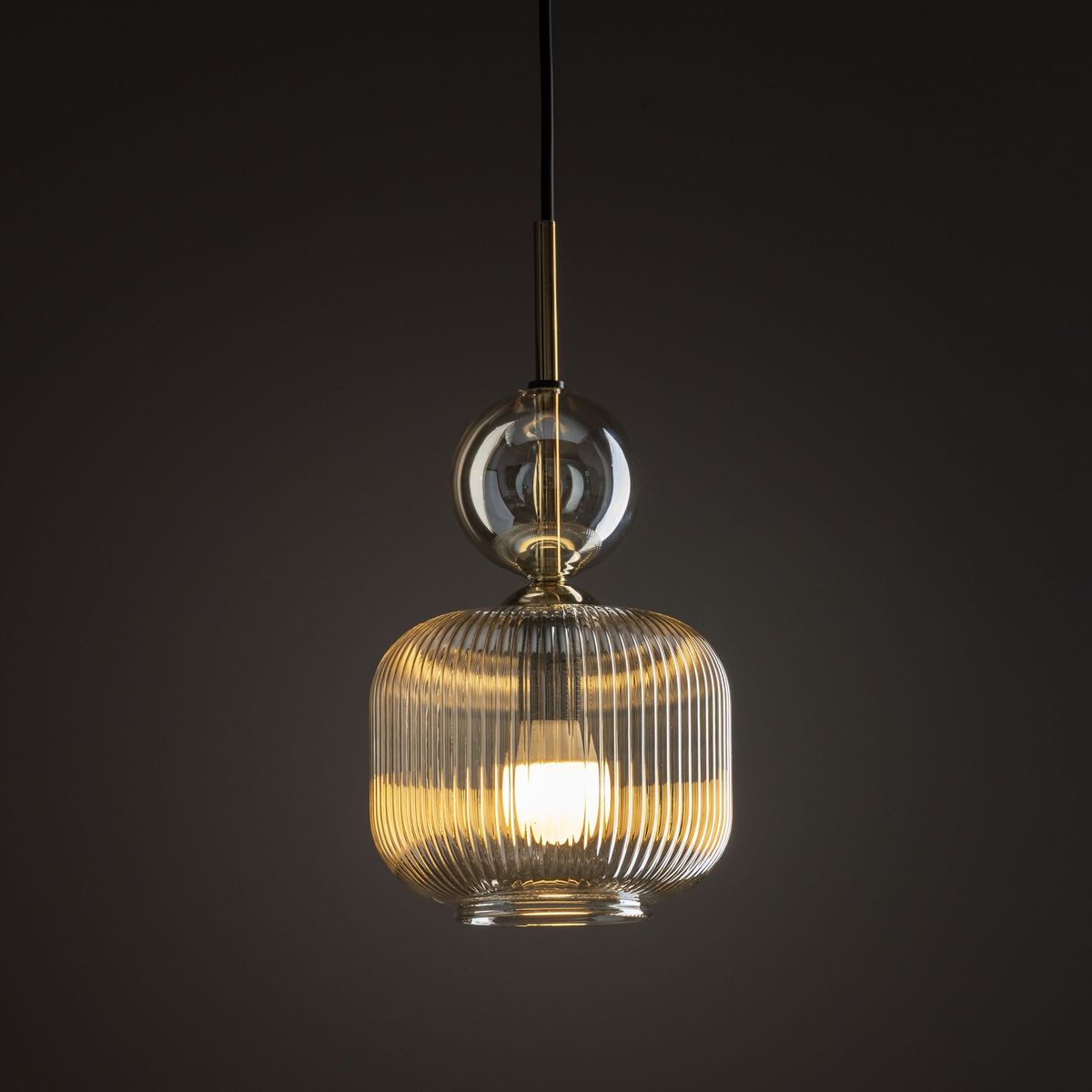 Lampa wisząca Sophia z kloszem w bursztynowym kolorze, z przezroczystym szklanym elementem u góry. Posiada jedno gniazdo E14. Zawieszona na czarnym przewodzie, w stylu nowoczesnym, z wyraźnym żłobieniem na kloszu. Tło ciemne.