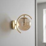 Złoty kinkiet El Dorado 1-punktowy z przezroczystą, okrągłą kloszem, zamontowany na ścianie. Klosz osadzony na metalowej konstrukcji z delikatnym łukiem, tworząc nowoczesny i elegancki wygląd. Tło w neutralnym kolorze.