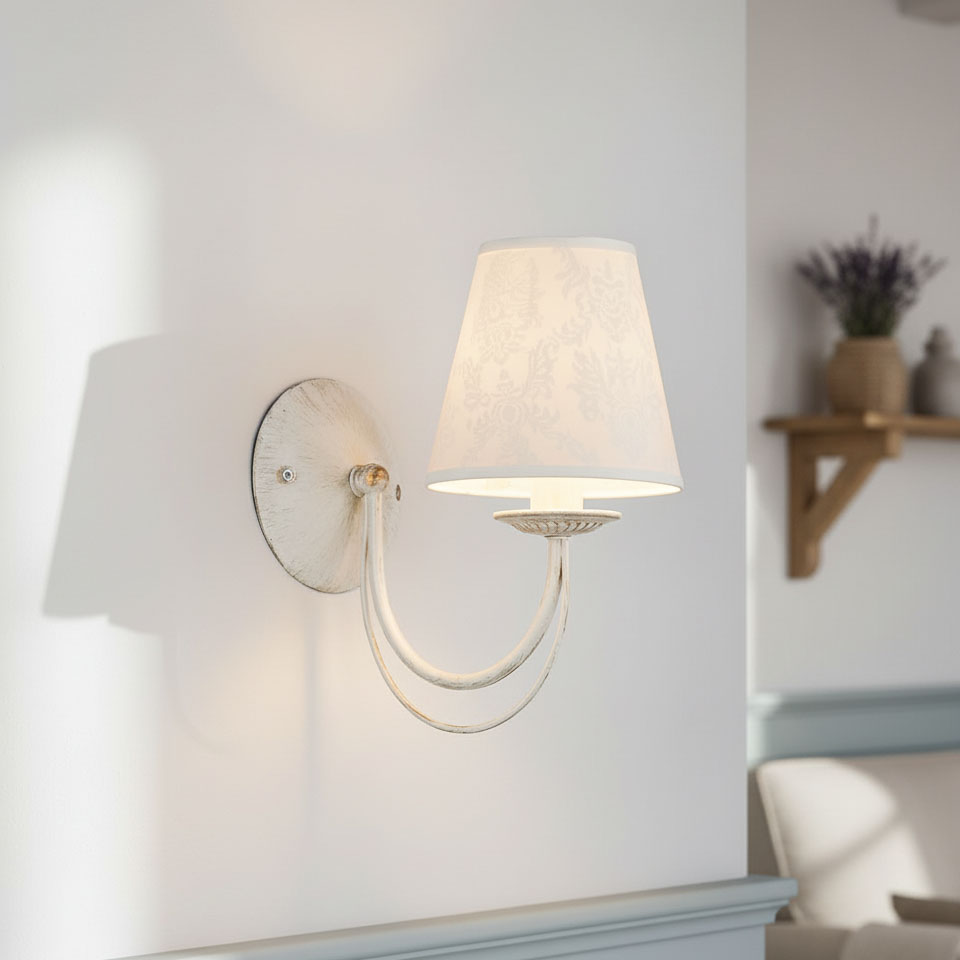 Kinkiet Irma 1-punktowy w stylu klasycznym, z białym abażurem i metalowym, lekko postarzałym uchwytem. Zawieszony na ścianie, z widocznym okrągłym mocowaniem. Tło jasne, minimalistyczne, z elementami aranżacyjnymi w salonie.