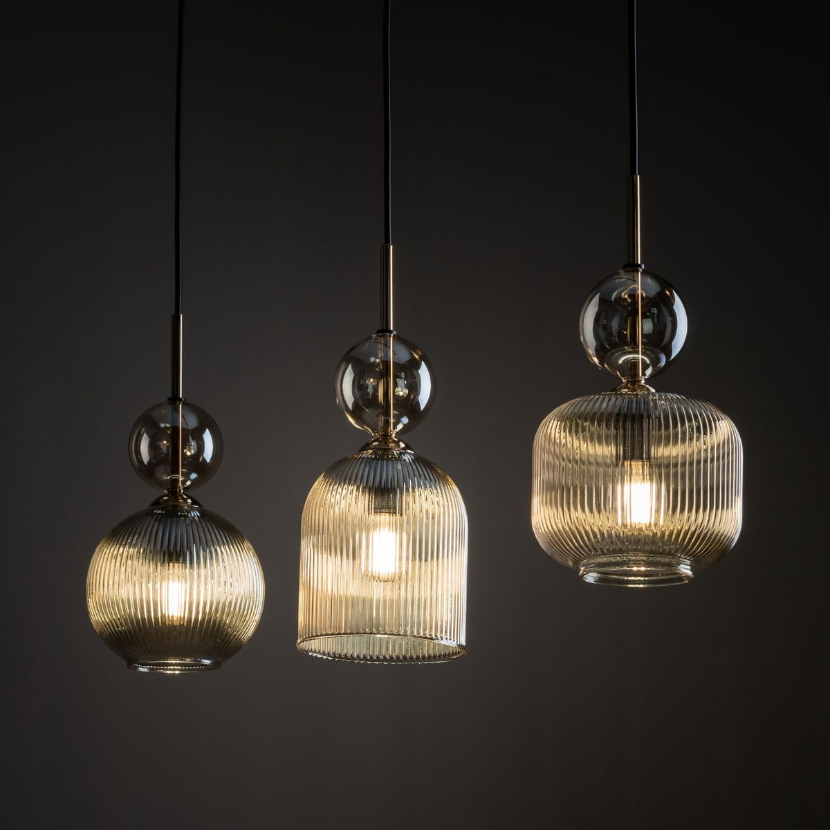 Lampa wisząca Sophia z trzema kloszami, wykonana z bursztynowego szkła z pionowymi żłobieniami. Klosze mają różne kształty: okrągły, stożkowaty i cylindryczny. Metalowe elementy w kolorze złotym, zawieszone na czarnych przewodach.