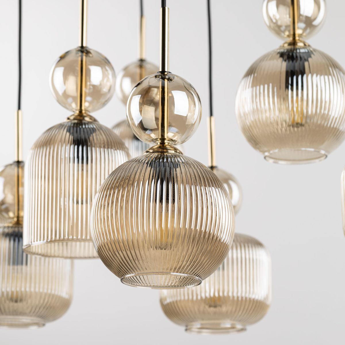 Lampa wisząca Sophia z 8 kloszami E14 w bursztynowym kolorze, z przezroczystymi, żłobionymi kloszami w kształcie cylindrów i kulistymi elementami, zawieszona na czarnych przewodach, umieszczona na jasnym tle.