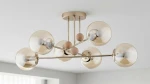 Złota lampa sufitowa Opty 6-punktowa z przezroczystymi kloszami, zawieszona w centralnej pozycji. Elementy drewniane oraz metalowe, z sześcioma żarówkami w kulistych kloszach. Tło jasne, minimalistyczne wnętrze.