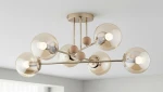 Lampa sufitowa Opty 6-punktowa w kolorze złotym z przezroczystymi kloszami. Posiada sześć kulistych źródeł światła oraz drewniane detale. Ujęcie z góry, produkt zamontowany na suficie. Tło jasne, minimalistyczne.
