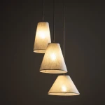 Lampa wisząca Coena Sabia z trzema kloszami w kształcie stożków, wykonanymi z beżowego materiału. Klosze zawieszone na cienkich przewodach, emitują ciepłe światło. Tło ciemne, co podkreśla delikatność lampy. Wymaga trzech żarówek E27.