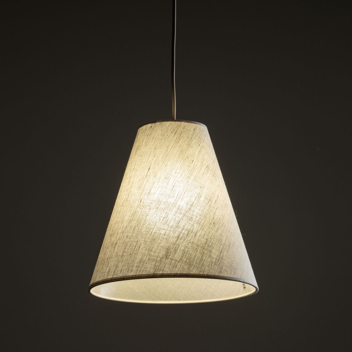 Beżowa lampa wisząca Coena Sabia z kloszem w kształcie stożka, zawieszona na cienkim przewodzie. Klosz wykonany z materiału, emituje miękkie światło. Ujęcie frontalne, na ciemnym tle, podkreślające teksturę i formę lampy.