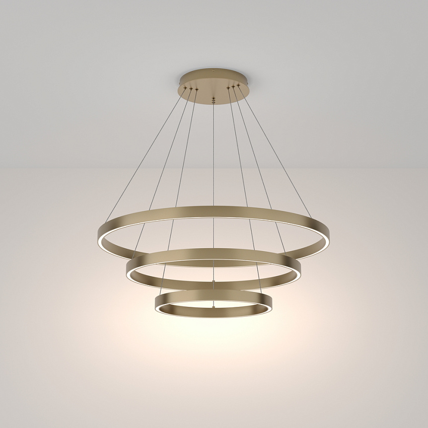 Lampa wisząca Rim 4000K, 3-punktowa, w kolorze mosiądzu. Składa się z trzech okrągłych pierścieni o różnych średnicach, zawieszonych na cienkich linkach. Źródło światła LED emituje jasne, białe światło, montowana na suficie na jednolitym tle.