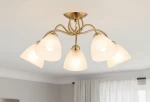 Lampa sufitowa Amarillo Patyna 5-punktowa w kolorze złotym z pięcioma alabastrowymi kloszami w kształcie stożka. Ujęcie z dołu, montowana na suficie, z eleganckimi, falującymi ramionami. Tło jasne, z zasłonami w oknie.
