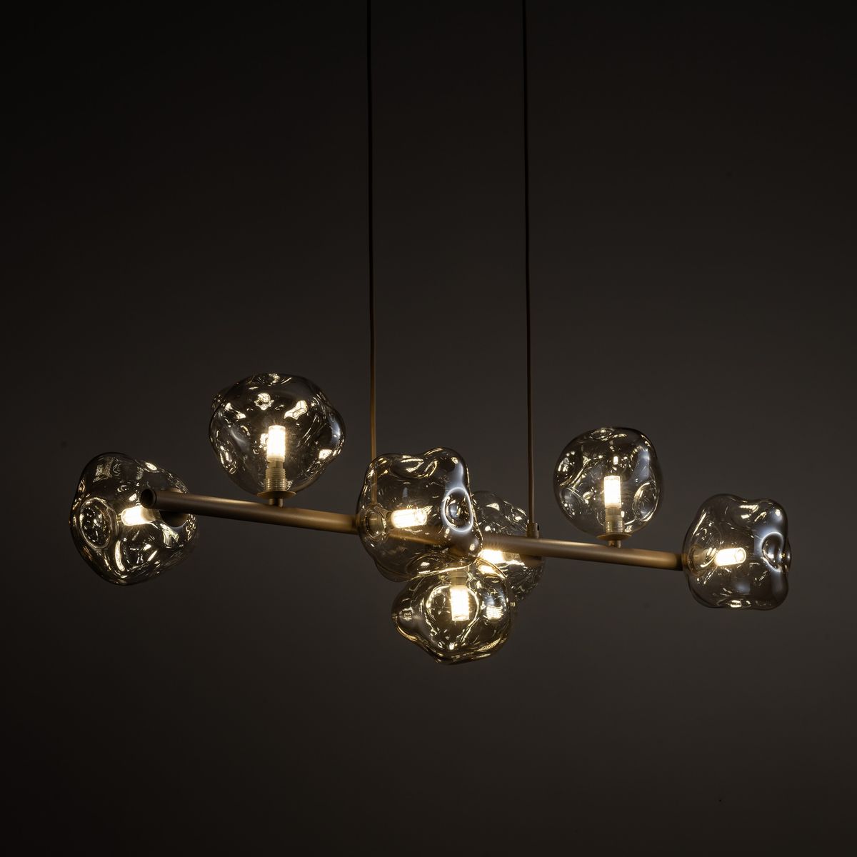 Lampa wisząca Lava Sabia w kolorze beżowym, z siedmioma szkłami o organicznych kształtach. Metalowa konstrukcja w neutralnym odcieniu, zawieszona na długich linkach, z widocznymi żarówkami G9. Tło ciemne, podkreślające elegancję lampy.